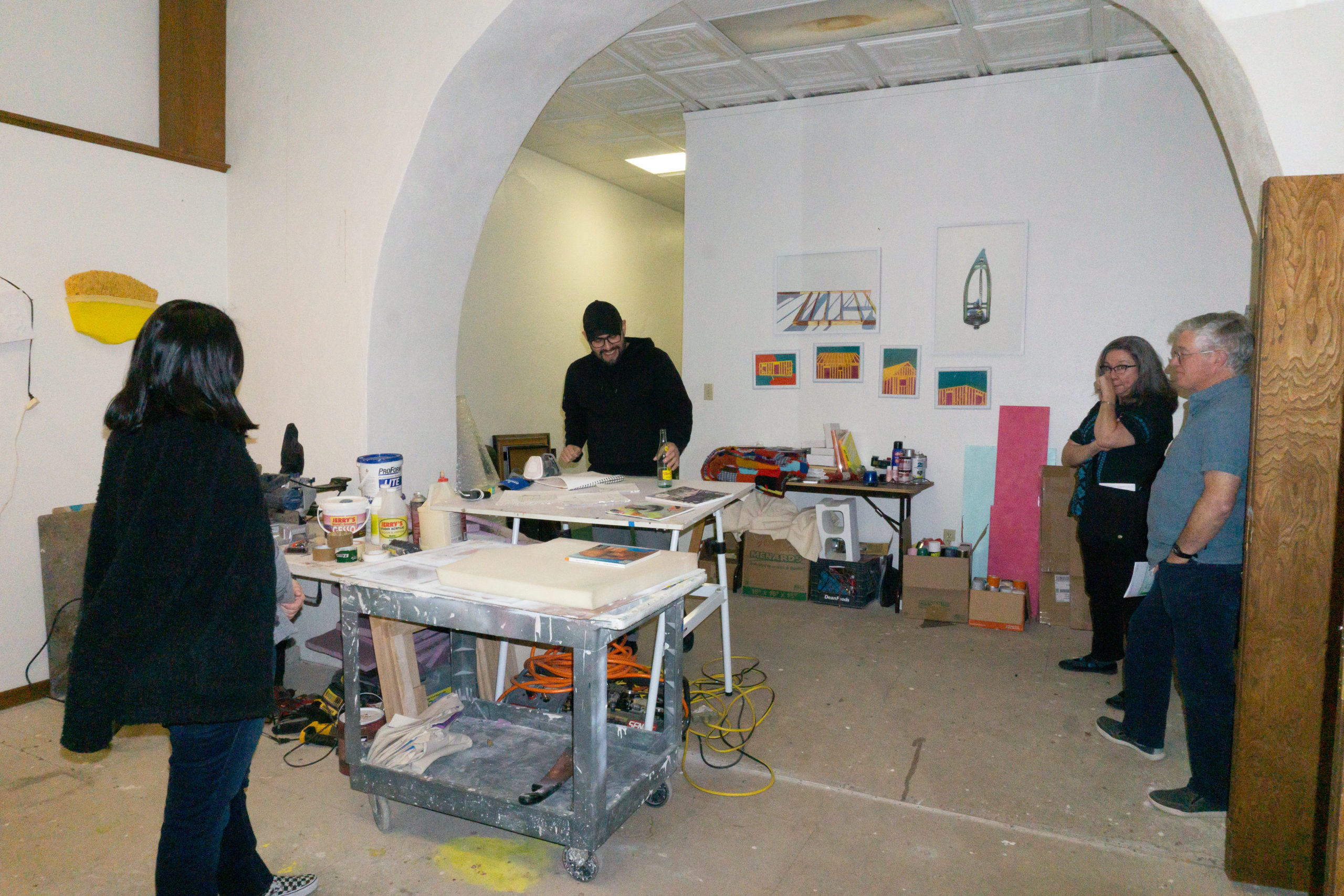 Michael Villarreal stands at a table in his art studio and speaks to a group.